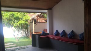 A covered outdoor seating area with a concrete floor, a low wall, and a view of a garden with trees and a thatched roof structure. - Backpackers Villa (Arugam Bay)
