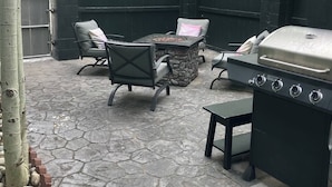An outdoor patio with stone flooring, a stone fire pit, a grill, and two chairs.
