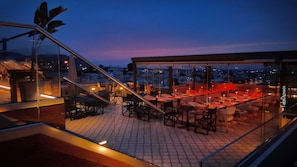 A rooftop restaurant with outdoor seating, a glass railing, and a city view at dusk. - Axel Beach Crete - Adults Only (Hersonissos)