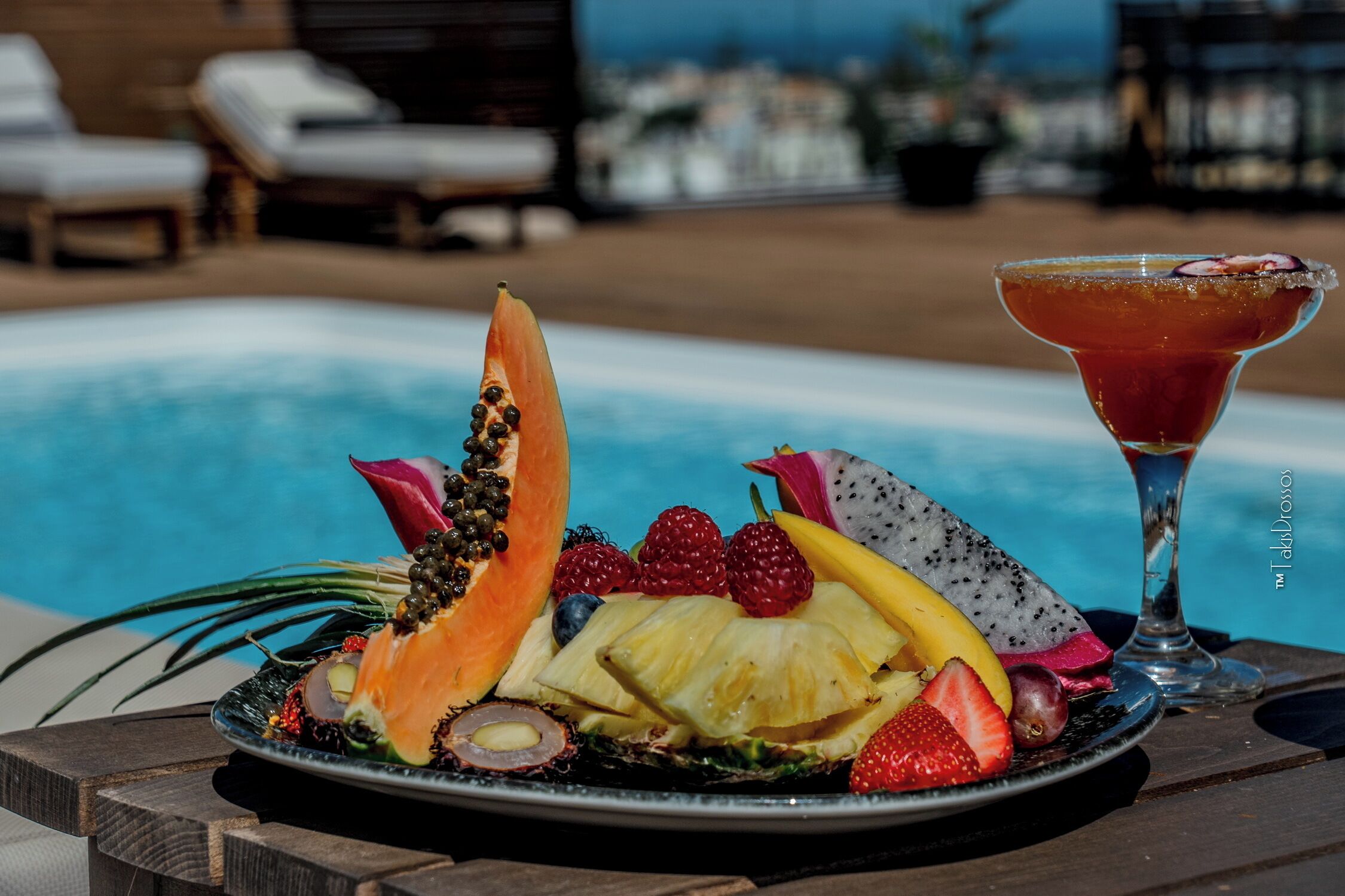 Una bandeja de frutas con una bebida tropical sobre una mesa junto a la piscina.