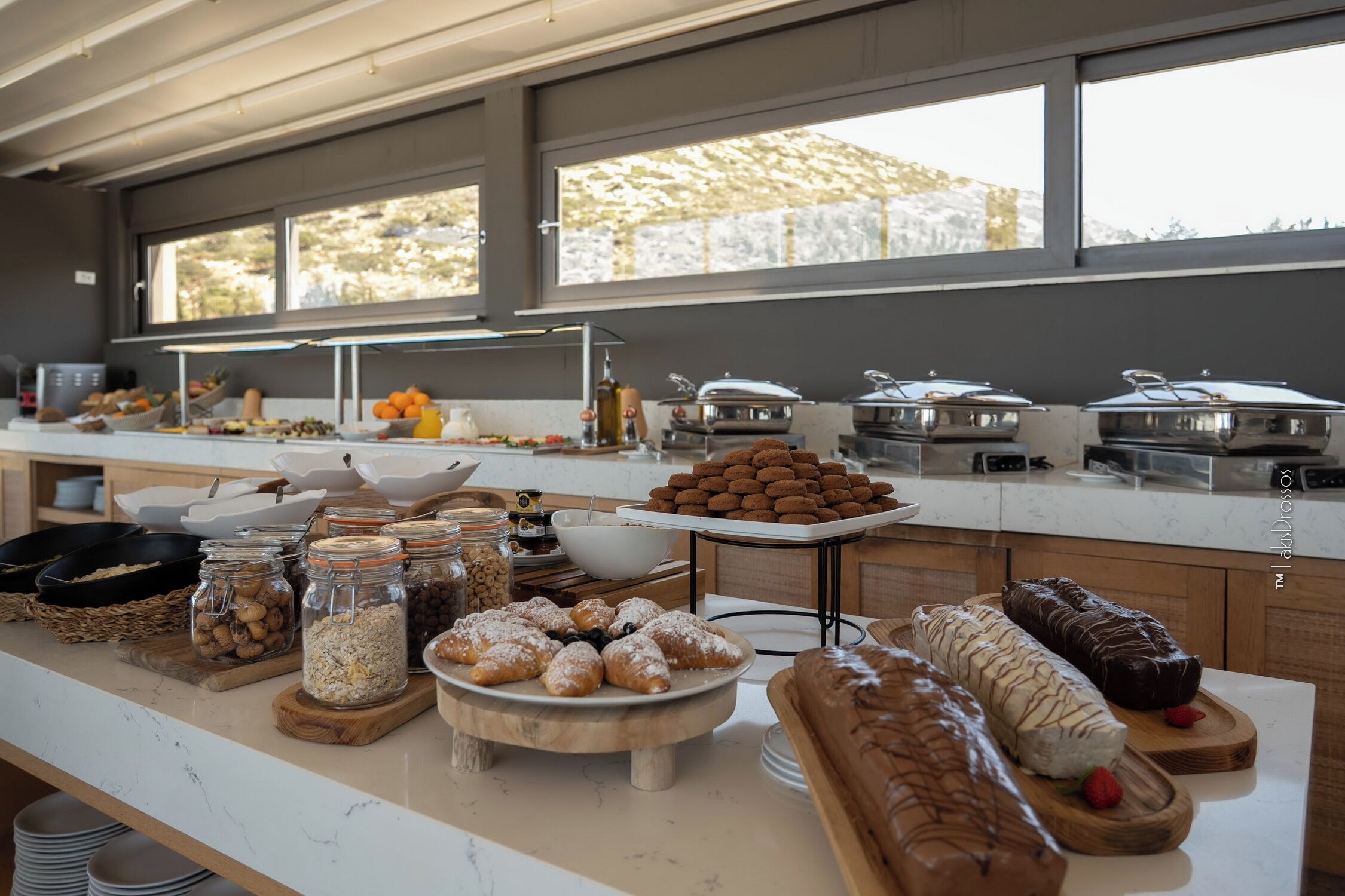 A buffet with various food items, including pastries and fruits, arranged on a counter with large windows in the background.
