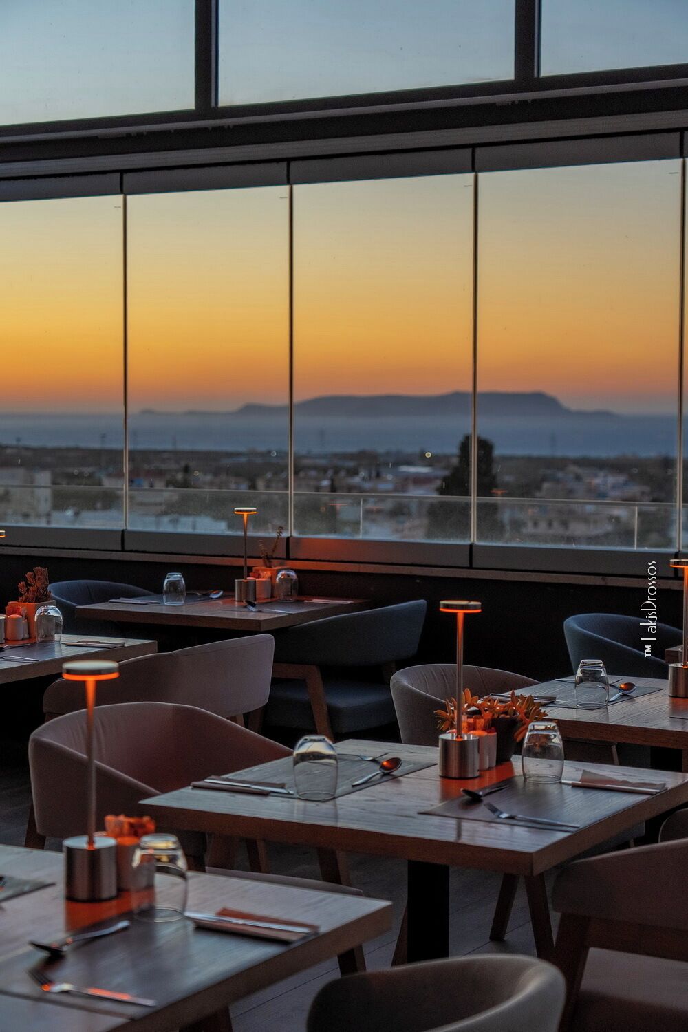 Een restaurant met tafels en stoelen, een uitzicht op de stad in de zonsondergang, en een groot raam.