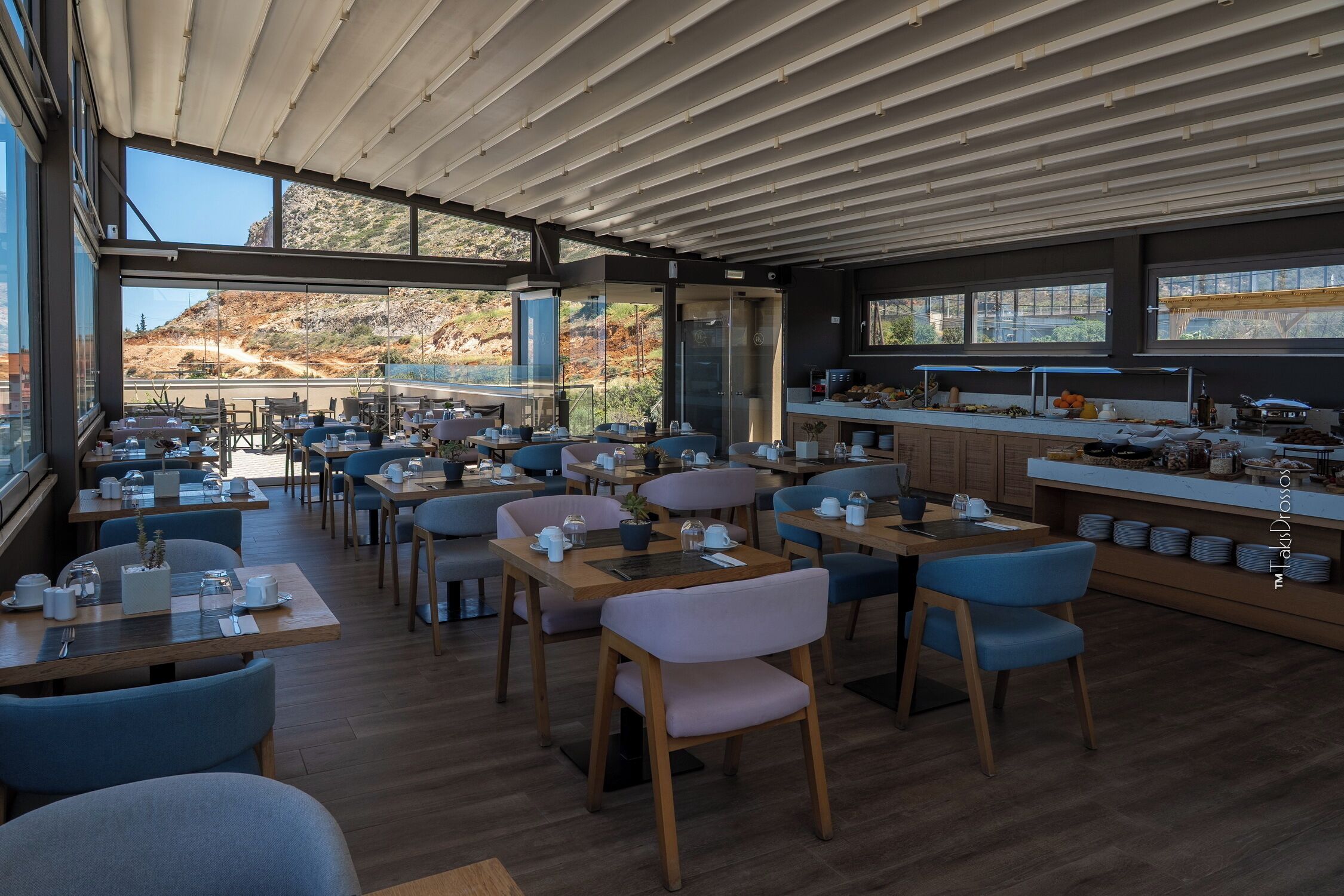 Un restaurante moderno con vistas a un paisaje montañoso, que cuenta con mesas de madera, sillas azules y una zona de cocina.