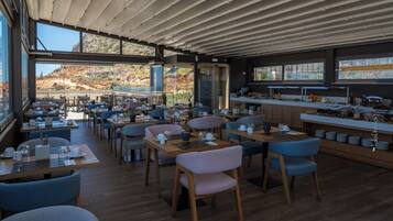 Un restaurante moderno con vistas a un paisaje montañoso, que cuenta con mesas de madera, sillas azules y una zona de cocina.