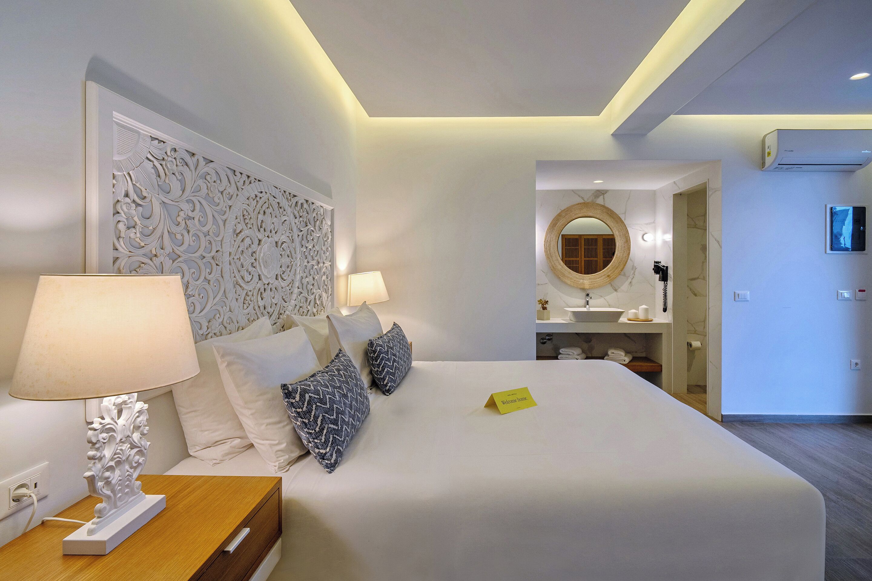 A hotel room with a large bed, two bedside lamps, a decorative headboard, a bedside table, a small bedside table, a mirror, a round mirror, a sink, a towel rack, and a television.