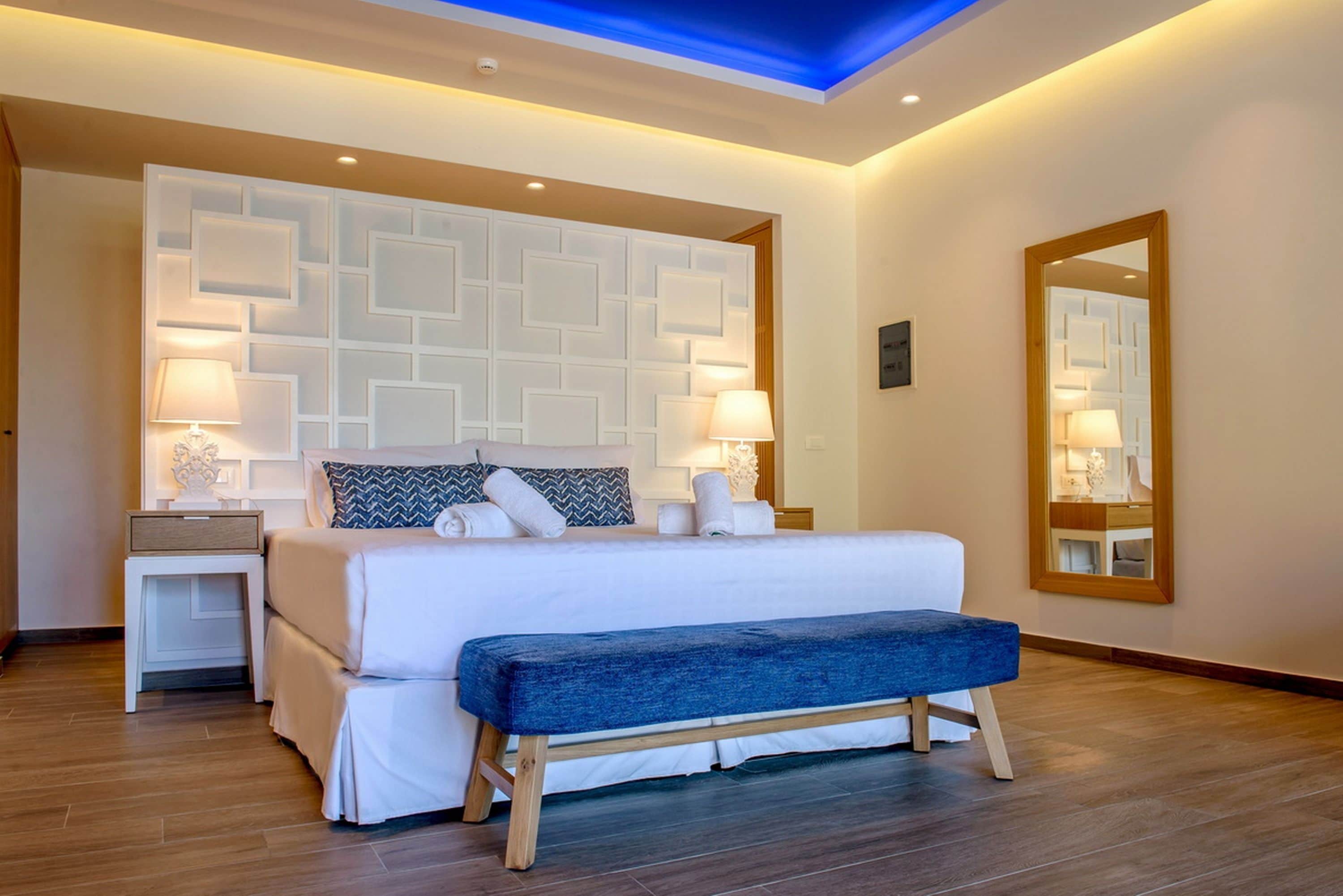 Quarto de hotel moderno com uma cama grande, abajures, um banco, um espelho e um painel de parede geométrico.