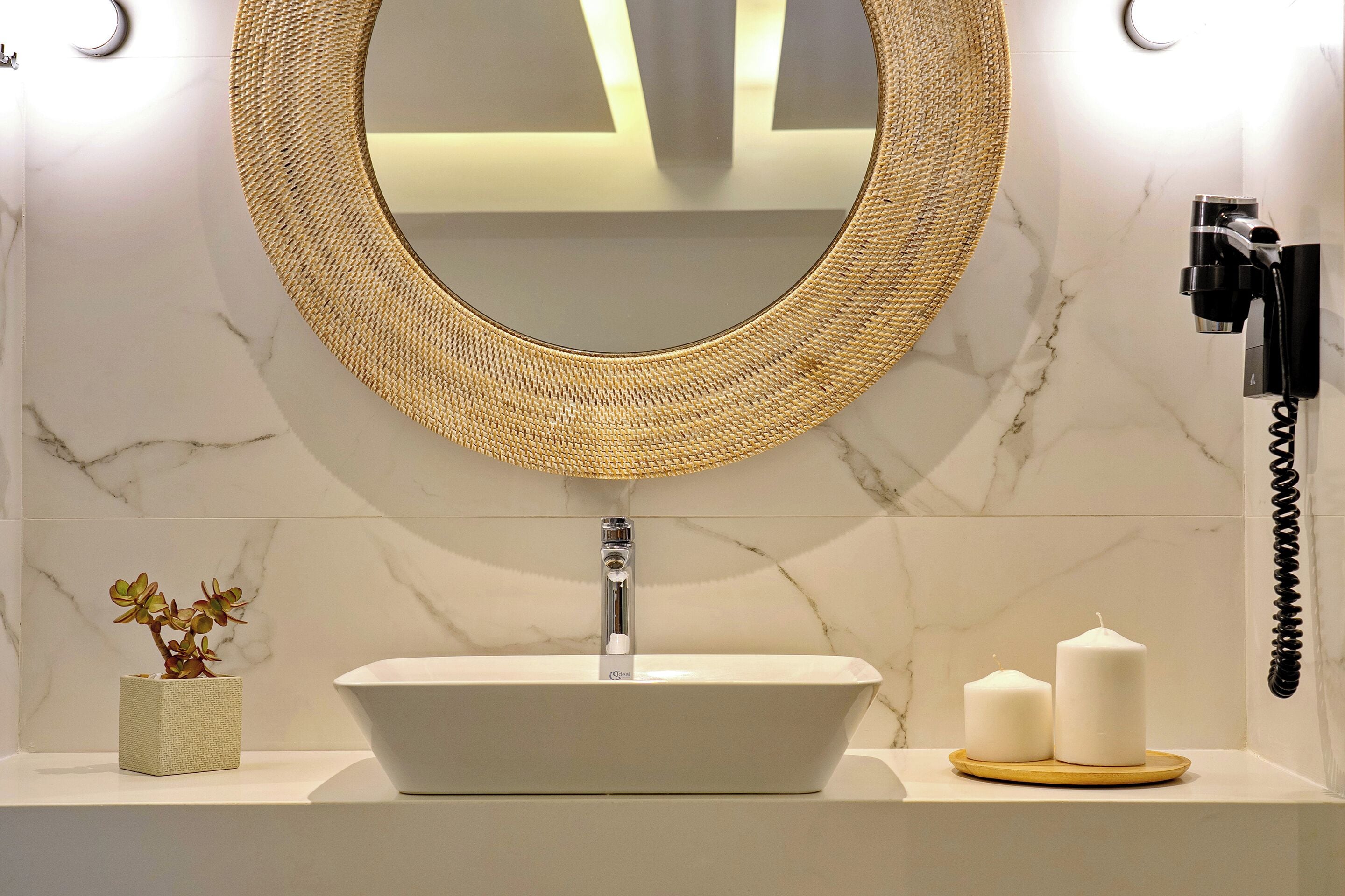A bathroom with a white sink, a round mirror with a woven frame, a small potted plant, and two candles on a tray.