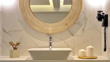 Een badkamer met een witte wasbak, een ronde spiegel met een gevlochten lijst, een kleine potplant en twee kaarsen op een dienblad.
