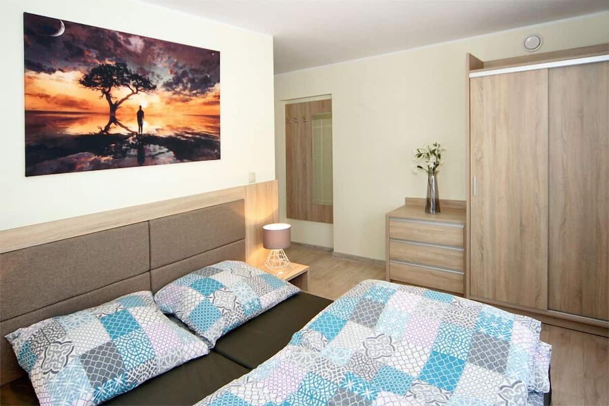 Ein Schlafzimmer mit Bett, Nachttisch, Lampe, Kleiderschrank und einem Bild von einem Baum am Wasser.