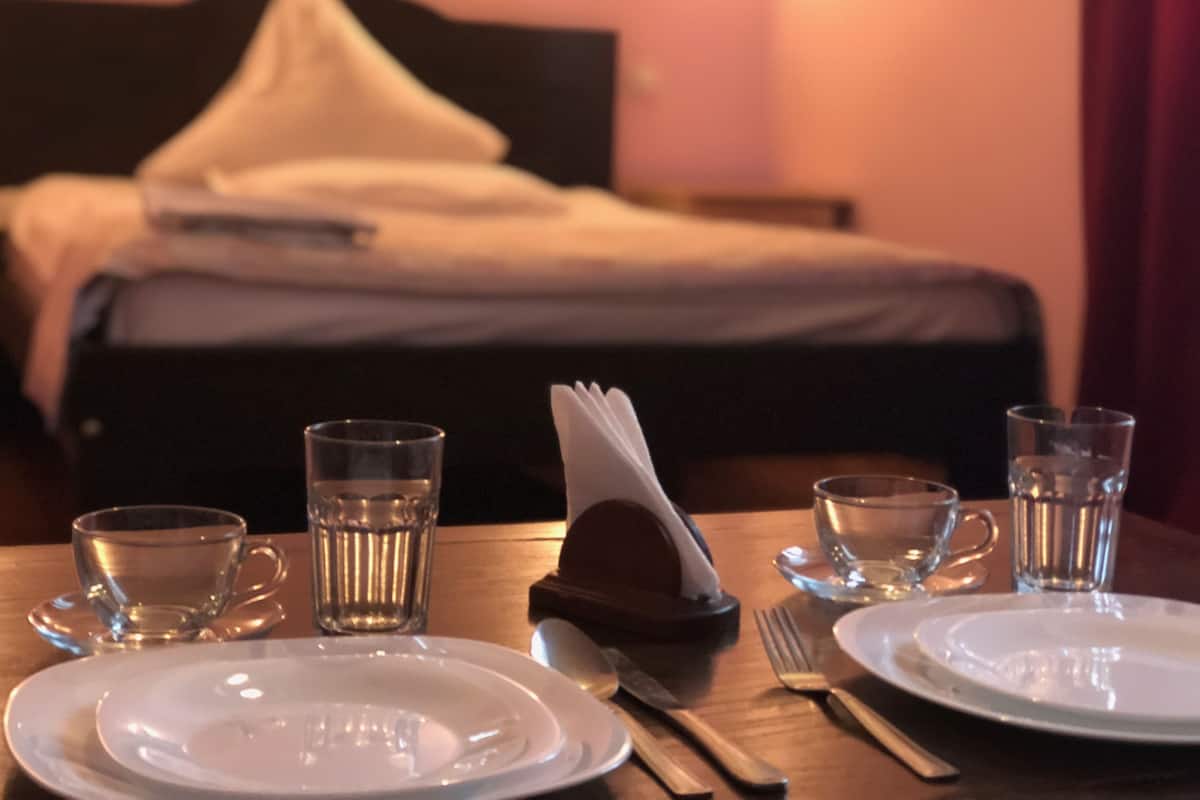 Una mesa de comedor bien puesta con platos, cubiertos y vasos, y una cama con un almohadón al fondo.