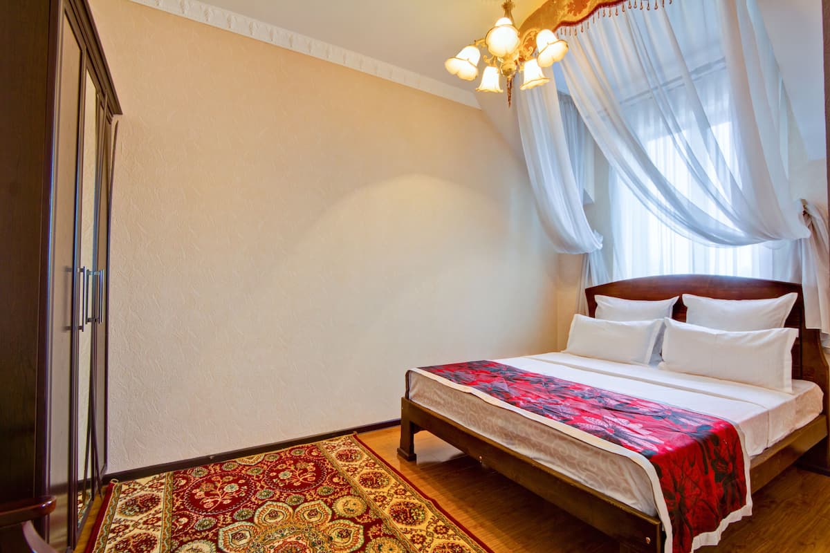 Un dormitorio con cama de madera, ropa de cama blanca, alfombra con estampado rojo y verde, candelabro y cortinas traslúcidas.