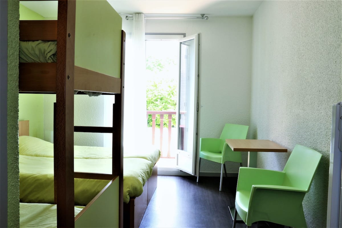 A small room with a bunk bed, a single chair, a table, and a door leading to a balcony.