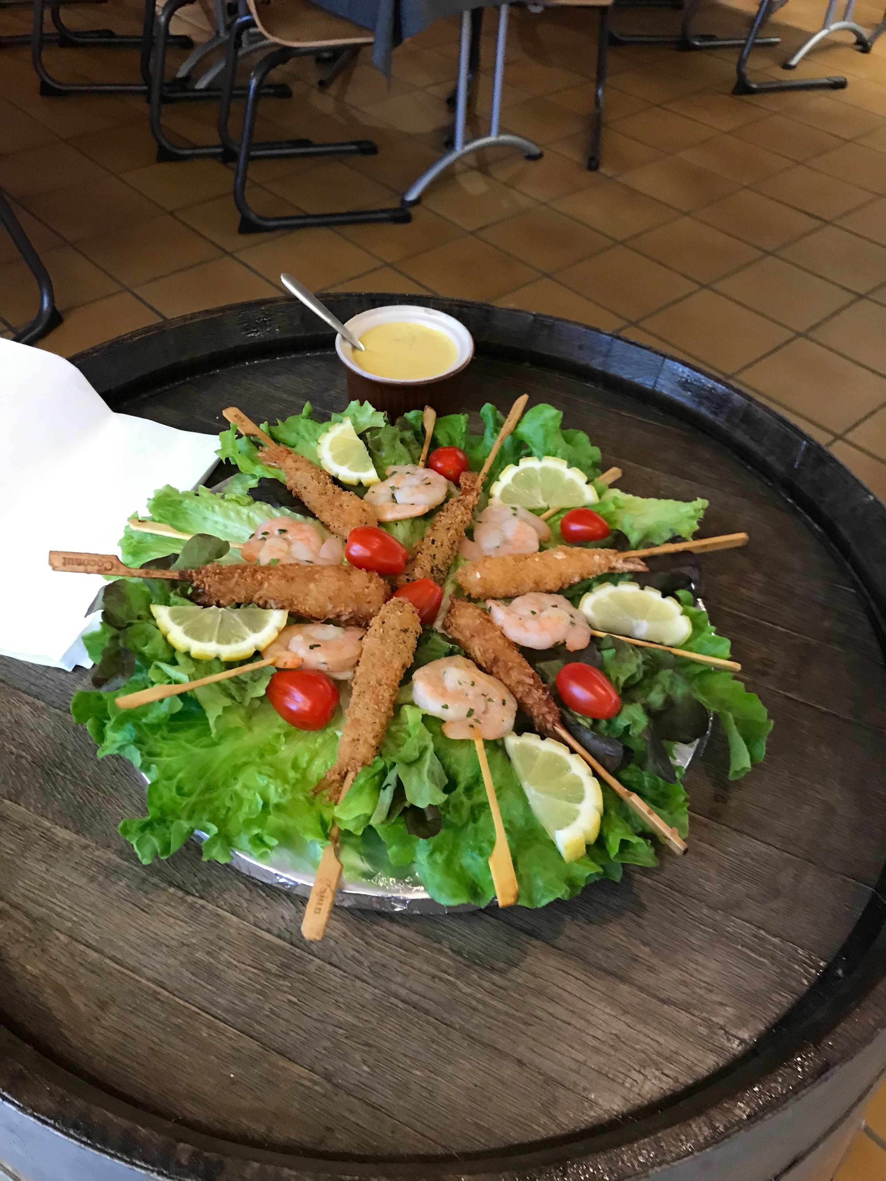 A round wooden table with a salad featuring shrimp skewers, lettuce, cherry tomatoes, and lemon slices.