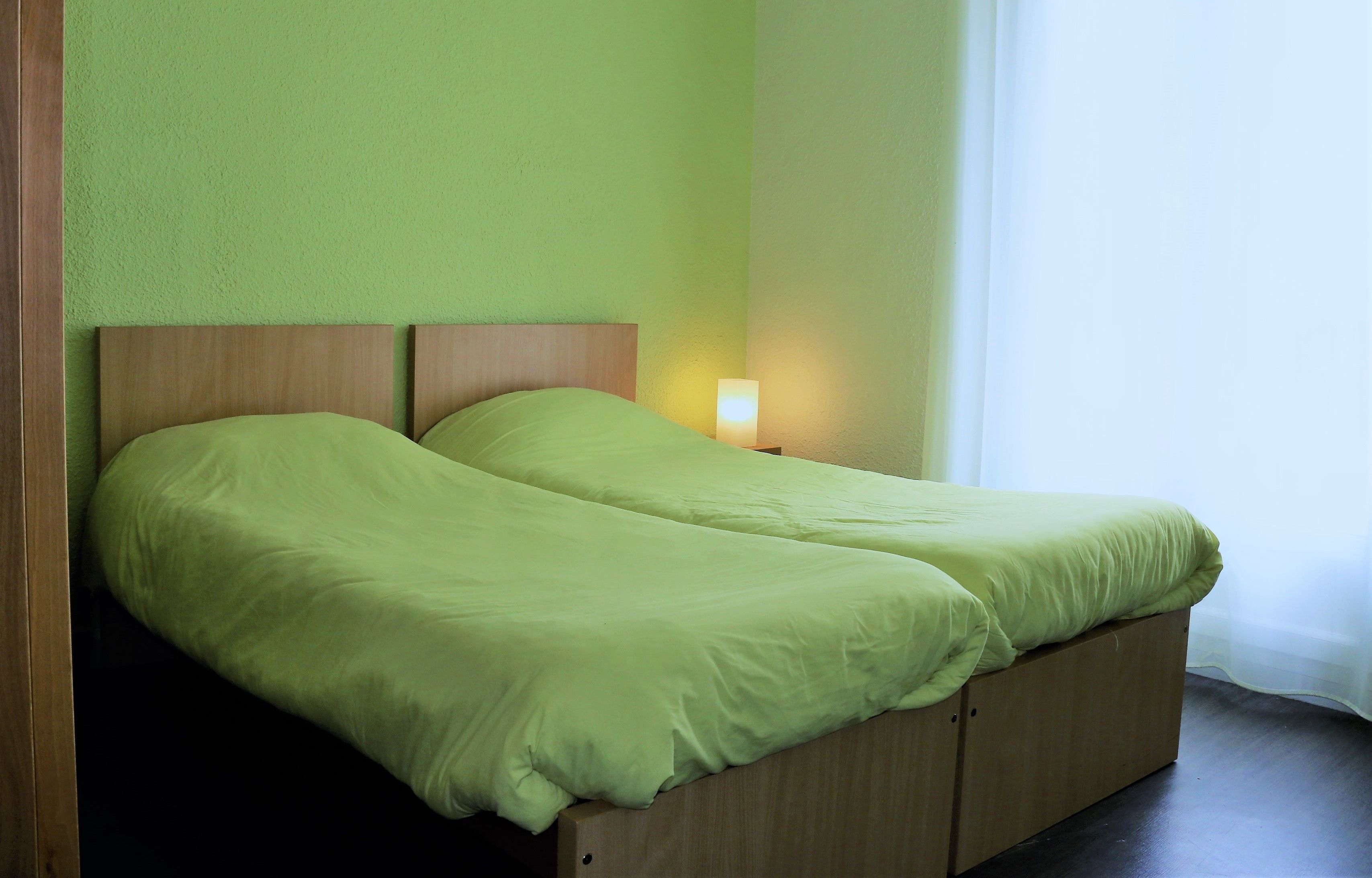 A double bed with a wooden headboard and a green bedspread. There is a bedside table with a lamp on it.