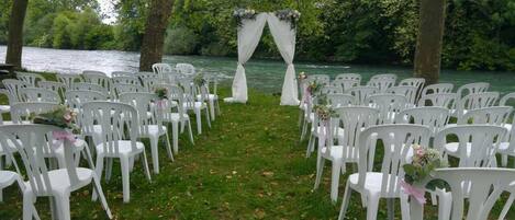 A riverside outdoor wedding setup with white plastic chairs arranged in rows, a white arch with floral decorations, and a grassy area.