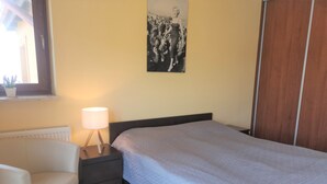 A bedroom with a bed, a chair, a nightstand with a lamp, a radiator, and a poster on the wall. - HoliApart Gdanska 52 (Krynica Morska)