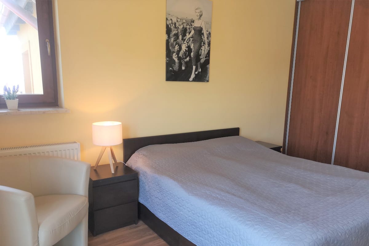 A bedroom with a bed, a chair, a nightstand with a lamp, a radiator, and a poster on the wall.