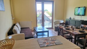 A cozy living room with a sofa, a dining table set for four, and a balcony with a view of buildings and trees. - HoliApart Gdanska 52 (Krynica Morska)