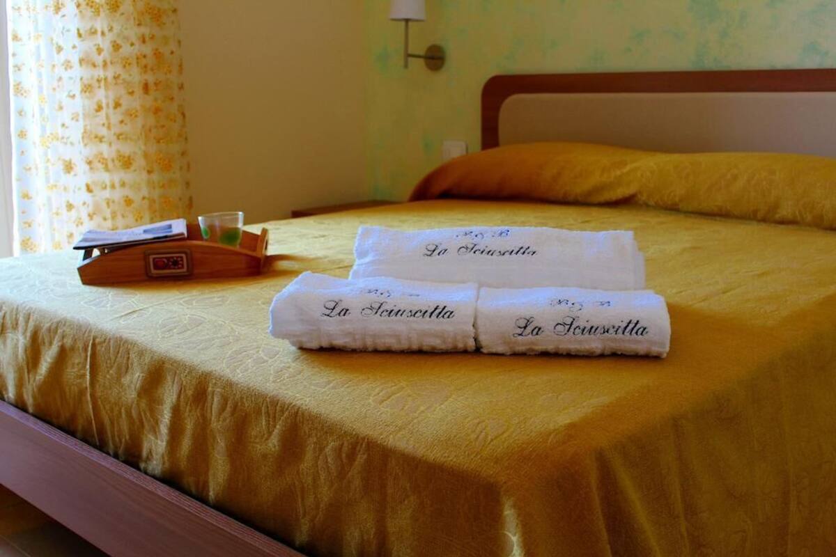 Un lit bien fait, recouvert d’une couverture jaune, deux serviettes pliées sur lesquelles est imprimé le nom « La Giustinia », un plateau en bois avec un verre d’eau et une lampe fixée au mur.