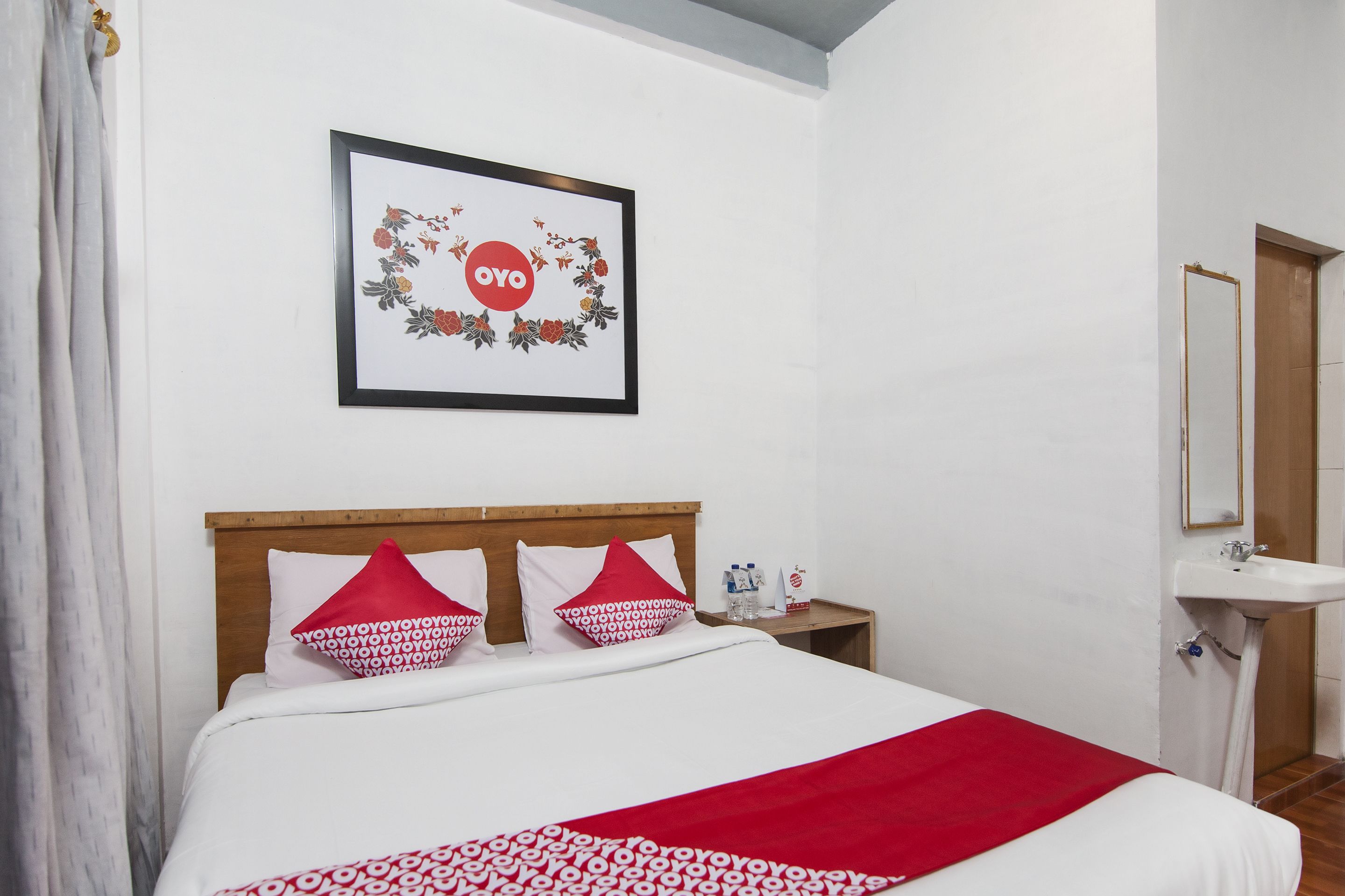 Ein ordentlich bezogenes Bett mit einer rot-weißen Decke, ein Holzkopfteil und zwei Kissen mit einem roten und weißen Muster. An der Wand über dem Bett hängt ein gerahmtes Bild, ein Nachttisch mit einer Flasche und einem Glas, und in der Ecke ein weißes Waschbecken.