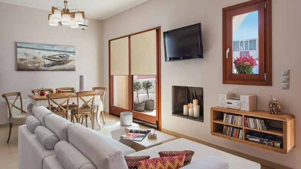 A modern living room with a sofa, a dining table, chairs, a TV, a bookshelf, and a window with a view. - Centro Houses (Santorini)