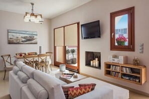 A modern living room with a sofa, a dining table, chairs, a TV, a bookshelf, and a window with a view. - Centro Houses (Santorini)