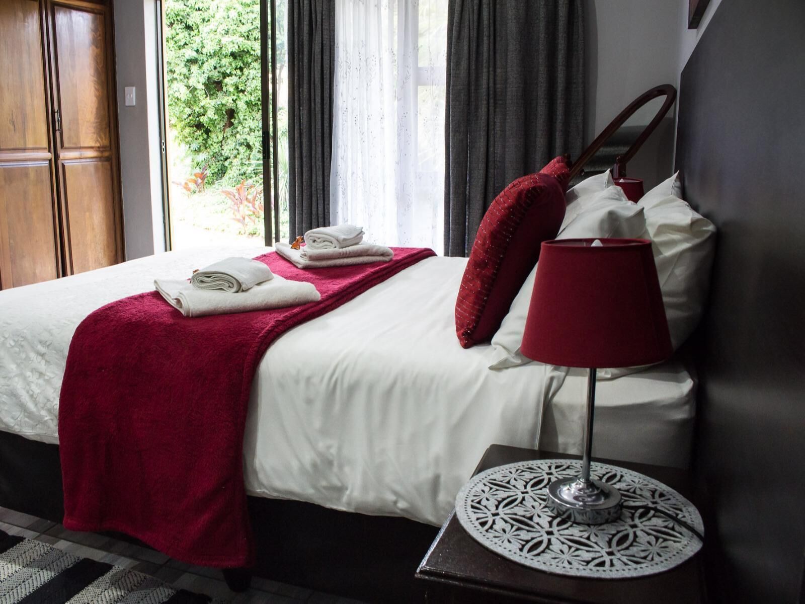 A neatly made bed with white linens and a red throw blanket, a red lamp on a bedside table, and a view of greenery outside the window.