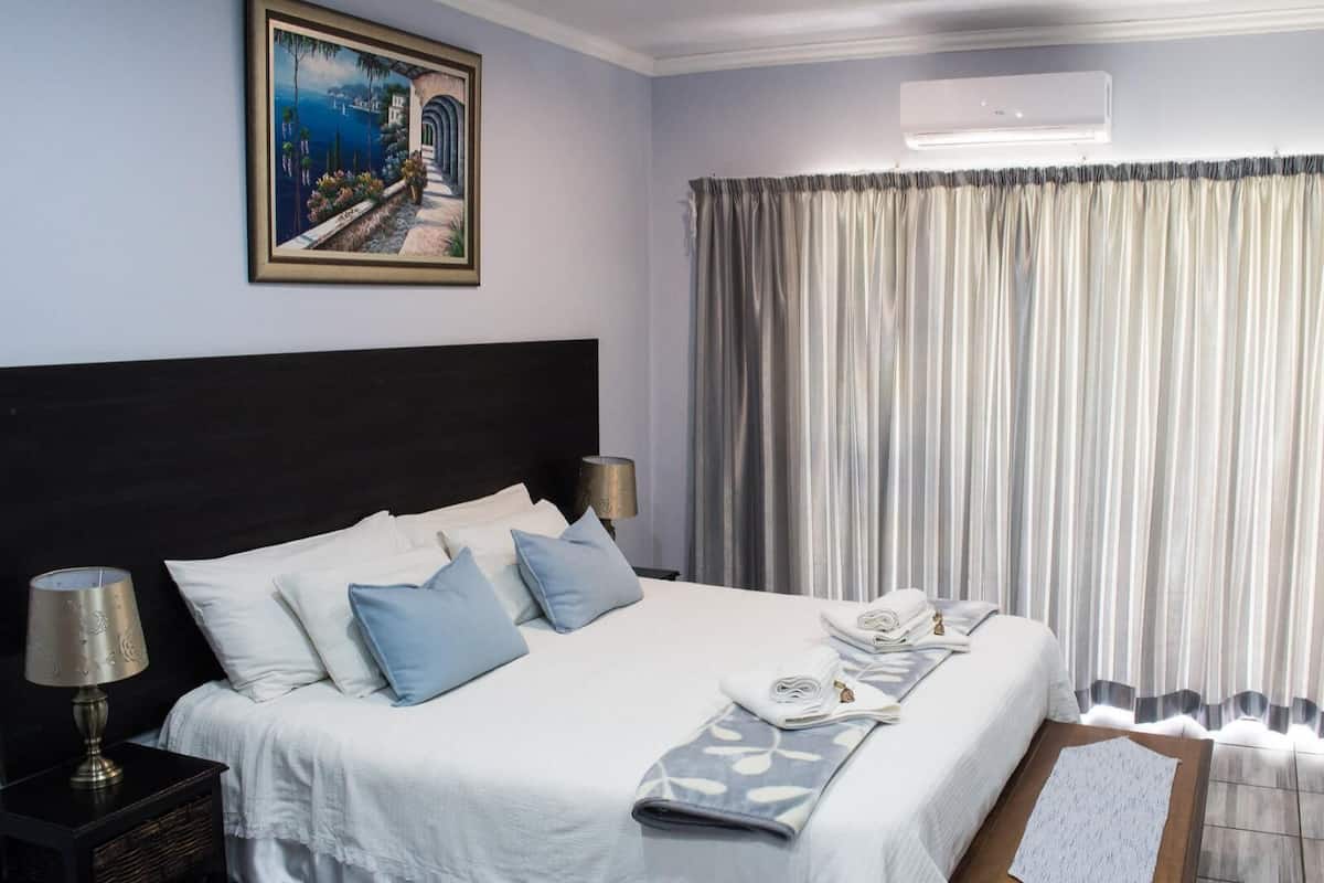 A neatly made bed with white and blue bedding, a lamp on a bedside table, and a painting on the wall.