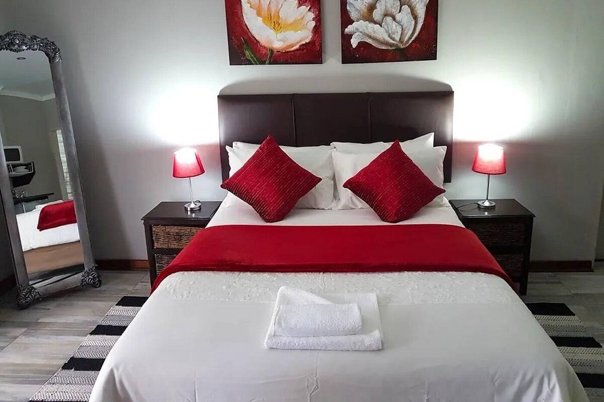 A neatly made bed with a red blanket and pillows, two bedside lamps, and two paintings of flowers on the wall.
