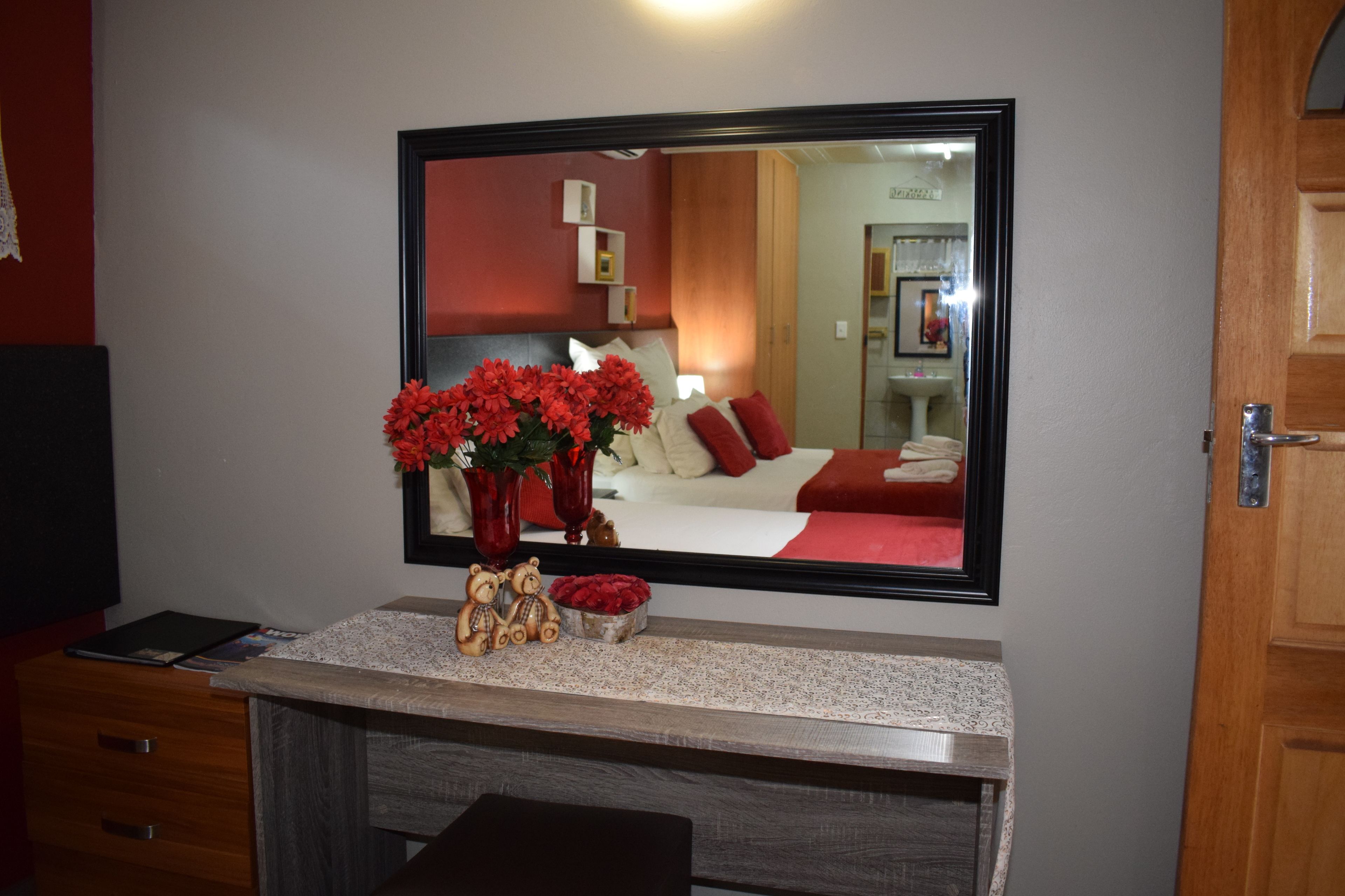 Quarto de hotel com um espelho grande, uma toucador com um vaso de flores vermelhas, uma cômoda de madeira e uma cama com roupa de cama vermelha.