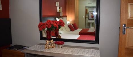 Une chambre d’hôtel avec un grand miroir, une coiffeuse ornée d’un vase de fleurs rouges, une commode en bois et un lit avec une literie rouge.