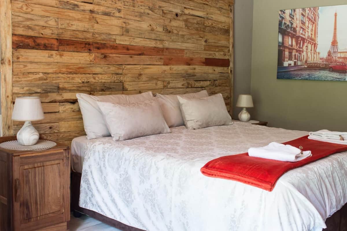 A bedroom with a wooden headboard, a bed with white bedding and a red throw blanket, two bedside lamps, and a painting of the Eiffel Tower on the wall.