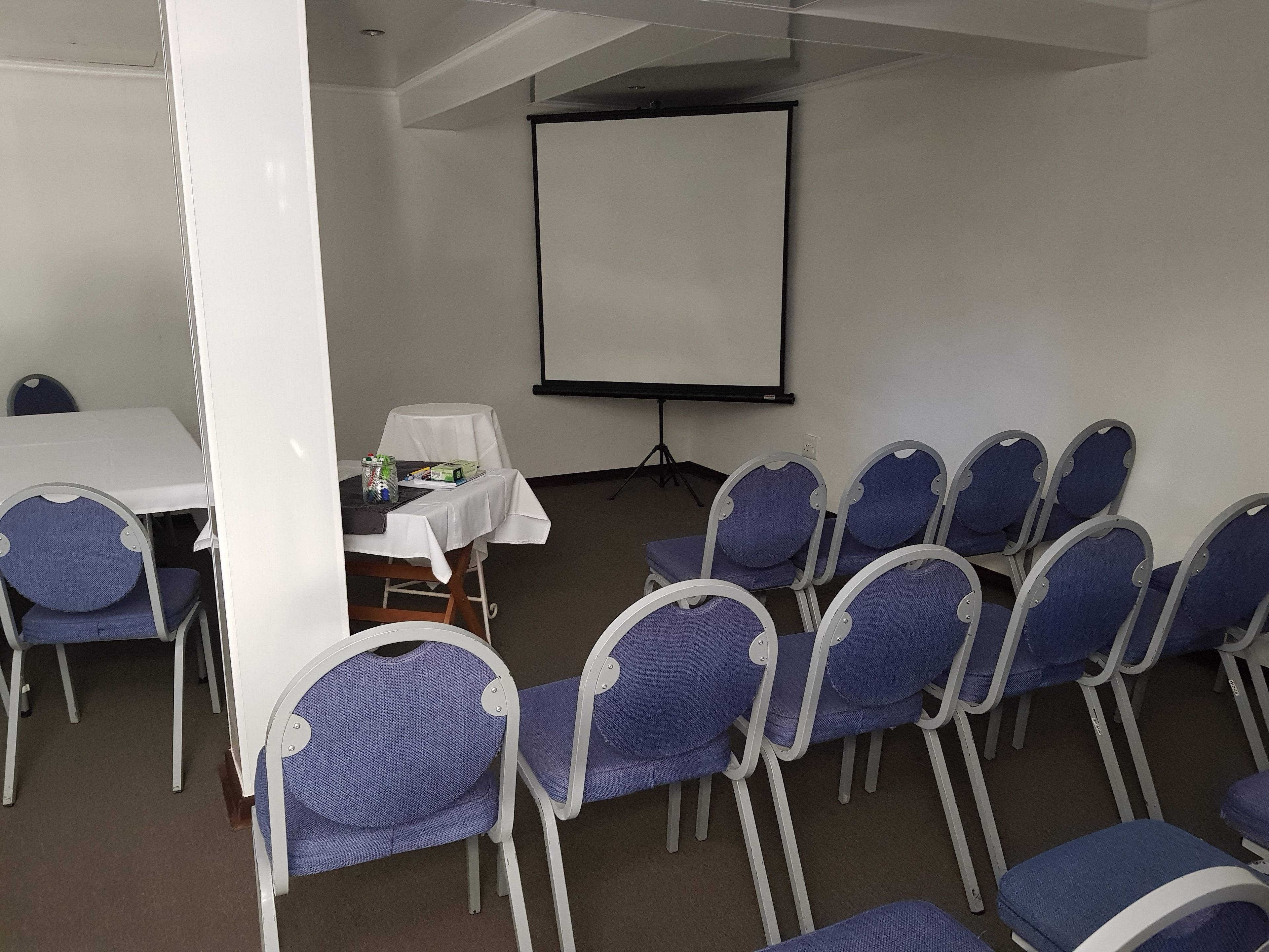 Un salón de conferencias con una mesa blanca, sillas, una pantalla de proyector y una pequeña mesa con mantel blanco y objetos encima.