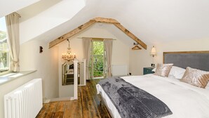 Um quarto com teto inclinado, uma cama com cobertor cinza, um candelabro, um radiador e uma janela com cortinas.