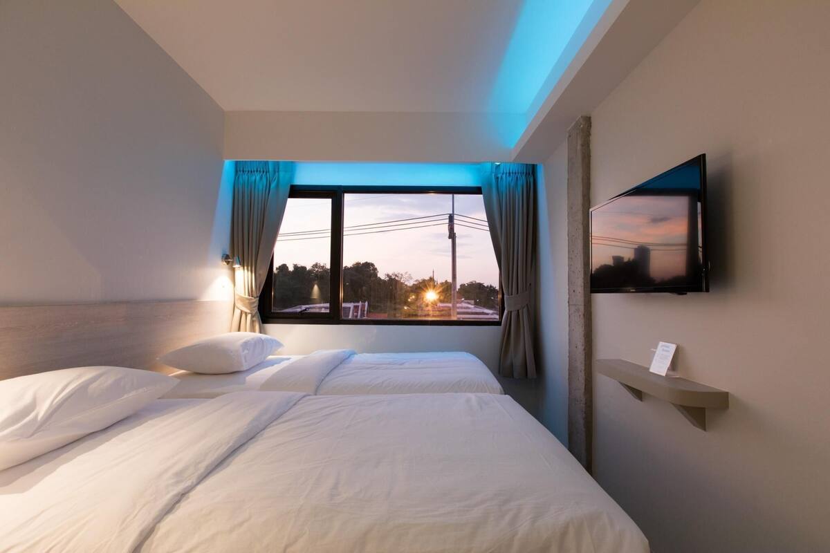 A hotel room with two beds, a TV mounted on the wall, a window with curtains, and a small shelf with a sign.