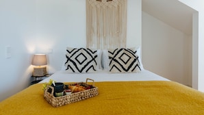 A bed with a yellow blanket, two patterned pillows, a macrame wall hanging, a bedside lamp, and a basket with a drink and snacks. - Boho Studio (Lisboa ciudad)