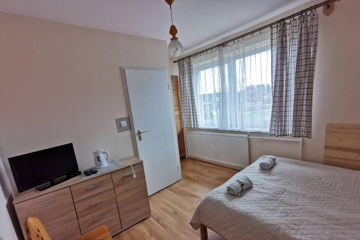 Ein Schlafzimmer mit Bett, Fernseher, Kommode und einem Fenster mit Vorhängen.