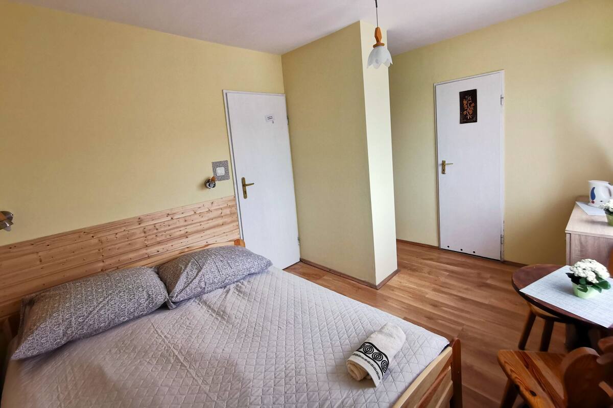 Ein Hotelzimmer mit einem Bett, einem Holzkopfstück, zwei Türen, einem kleinen Tisch mit Tasse und Blumen sowie einer Pendelleuchte.