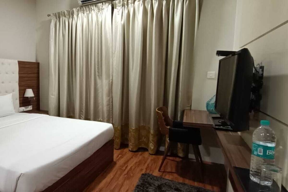 A hotel room with a large bed, a wooden headboard, a nightstand with a lamp, a desk with a chair, a television, and a curtain.