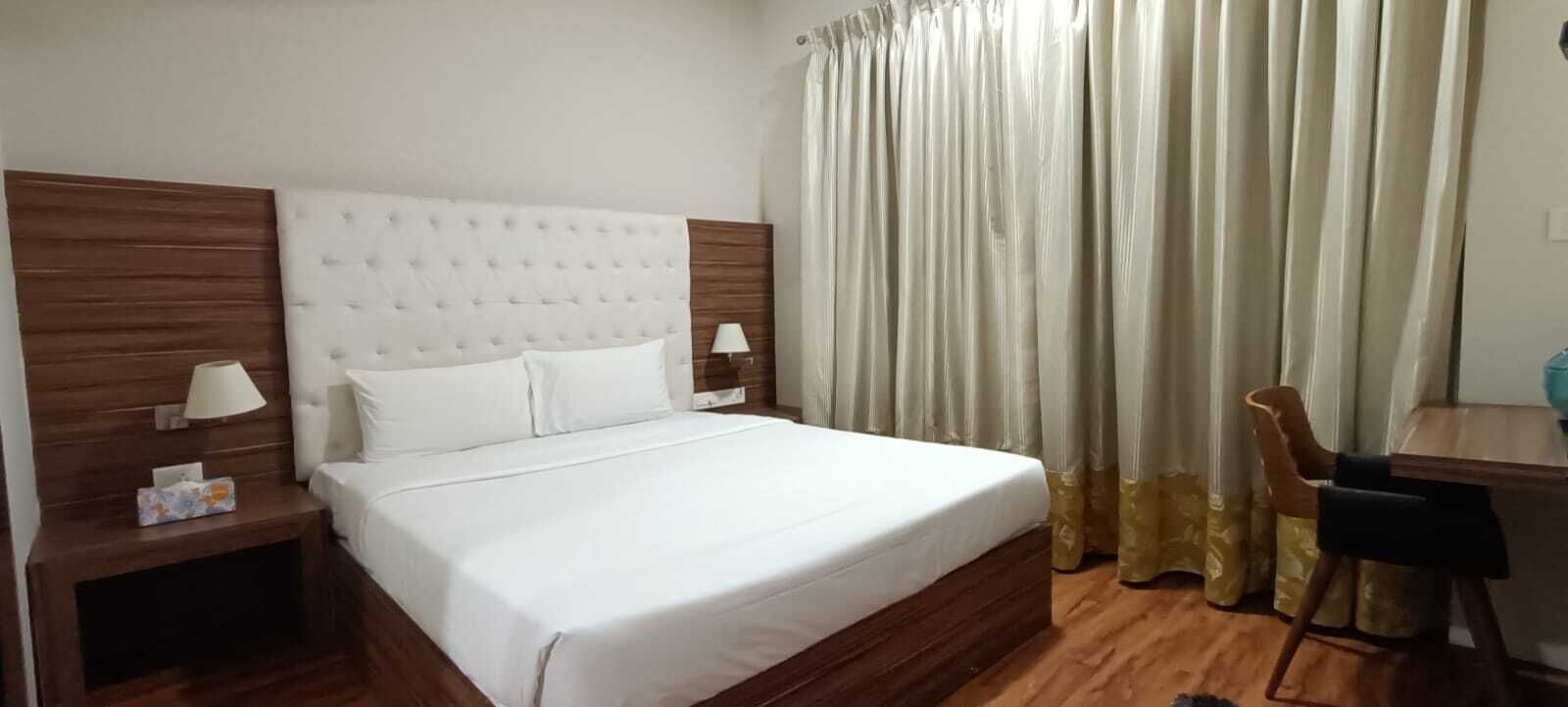 A neatly made bed with a headboard, two bedside tables with lamps, a chair, and a curtain.
