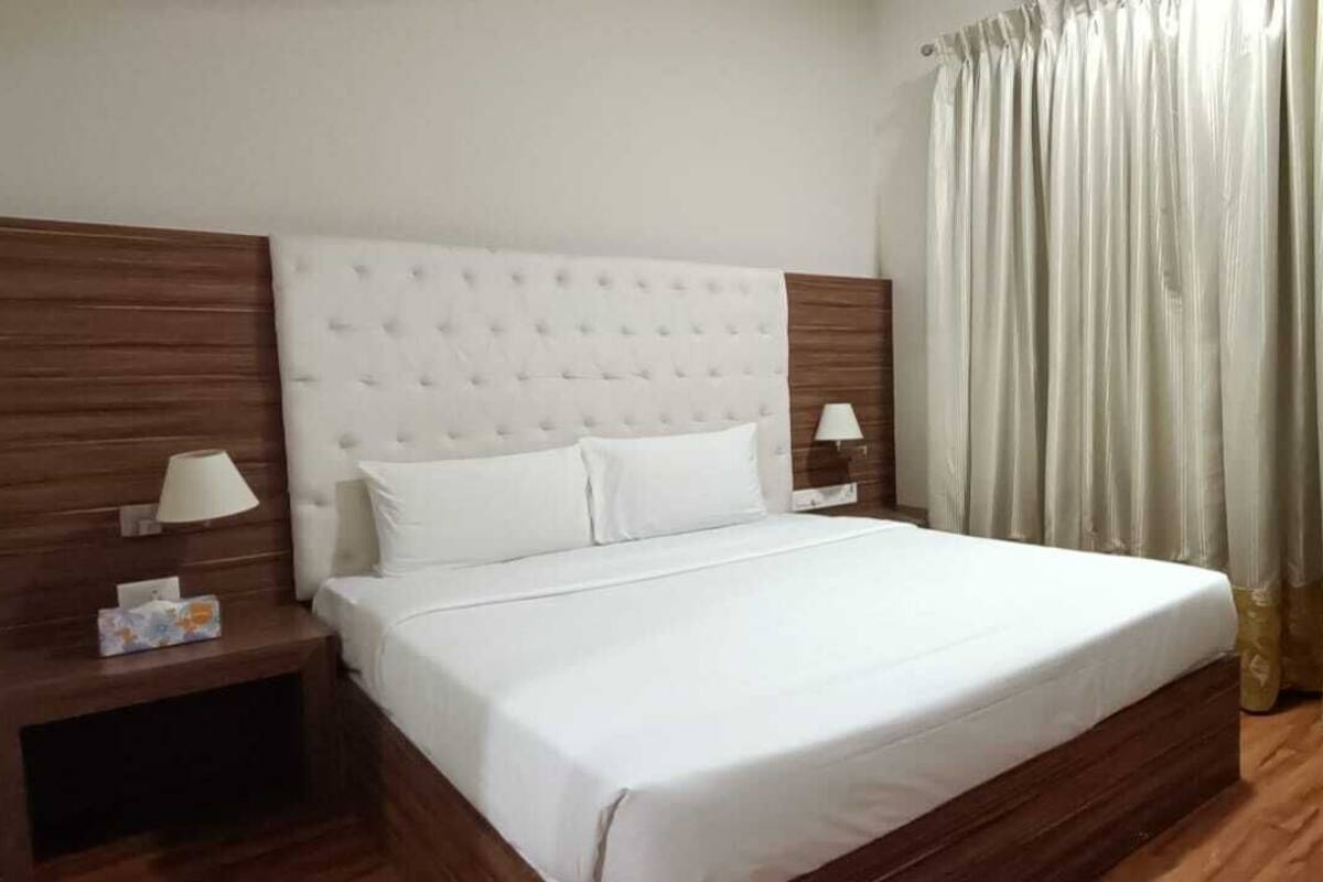 A neatly made bed with a headboard, two bedside tables with lamps, a chair, and a curtain.