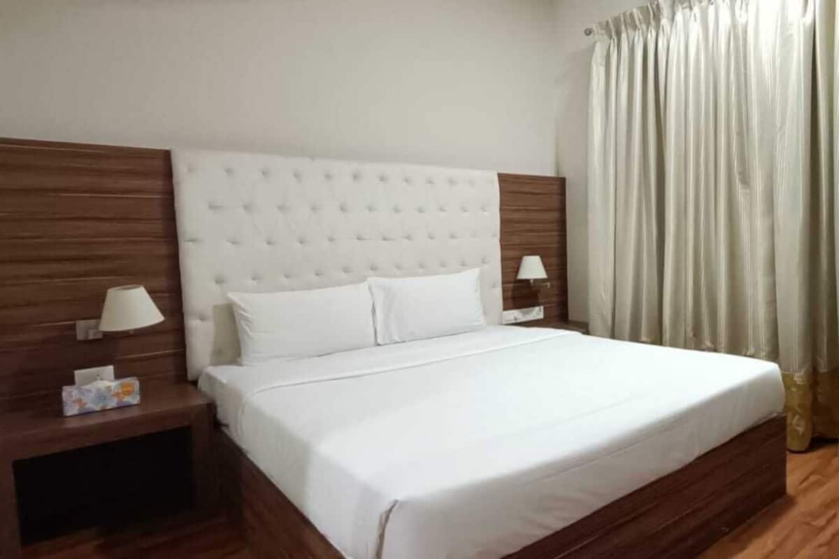 A neatly made bed with a headboard, two bedside tables with lamps, a chair, and a curtain.