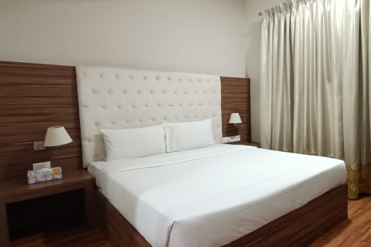 A neatly made bed with a headboard, two bedside tables with lamps, a chair, and a curtain.