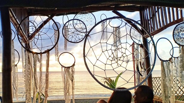 A couple relaxing in a hammock under a thatched roof, overlooking a beach with dreamcatchers hanging above. - Pacific Buddies - Campsite (Dingalan)