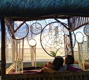 A couple relaxing in a hammock under a thatched roof, overlooking a beach with dreamcatchers hanging above. - Pacific Buddies - Campsite (Dingalan)