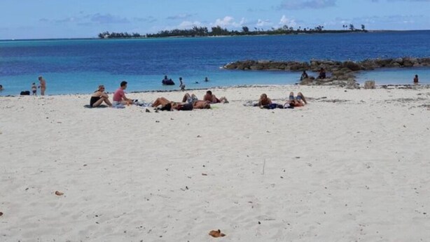 Uma praia com pessoas tomando sol e nadando, céu claro e uma ilha ao longe.