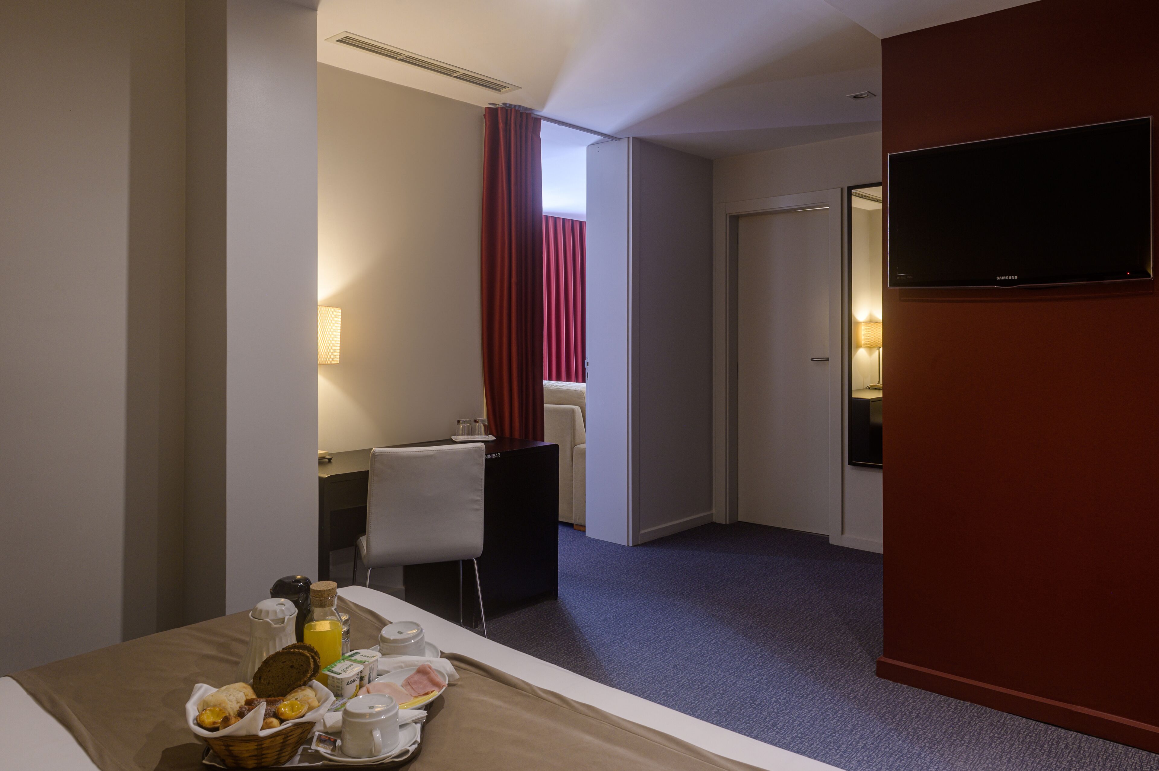 A hotel room with a bed, breakfast tray, desk, chair, television, and a door leading to another room.
