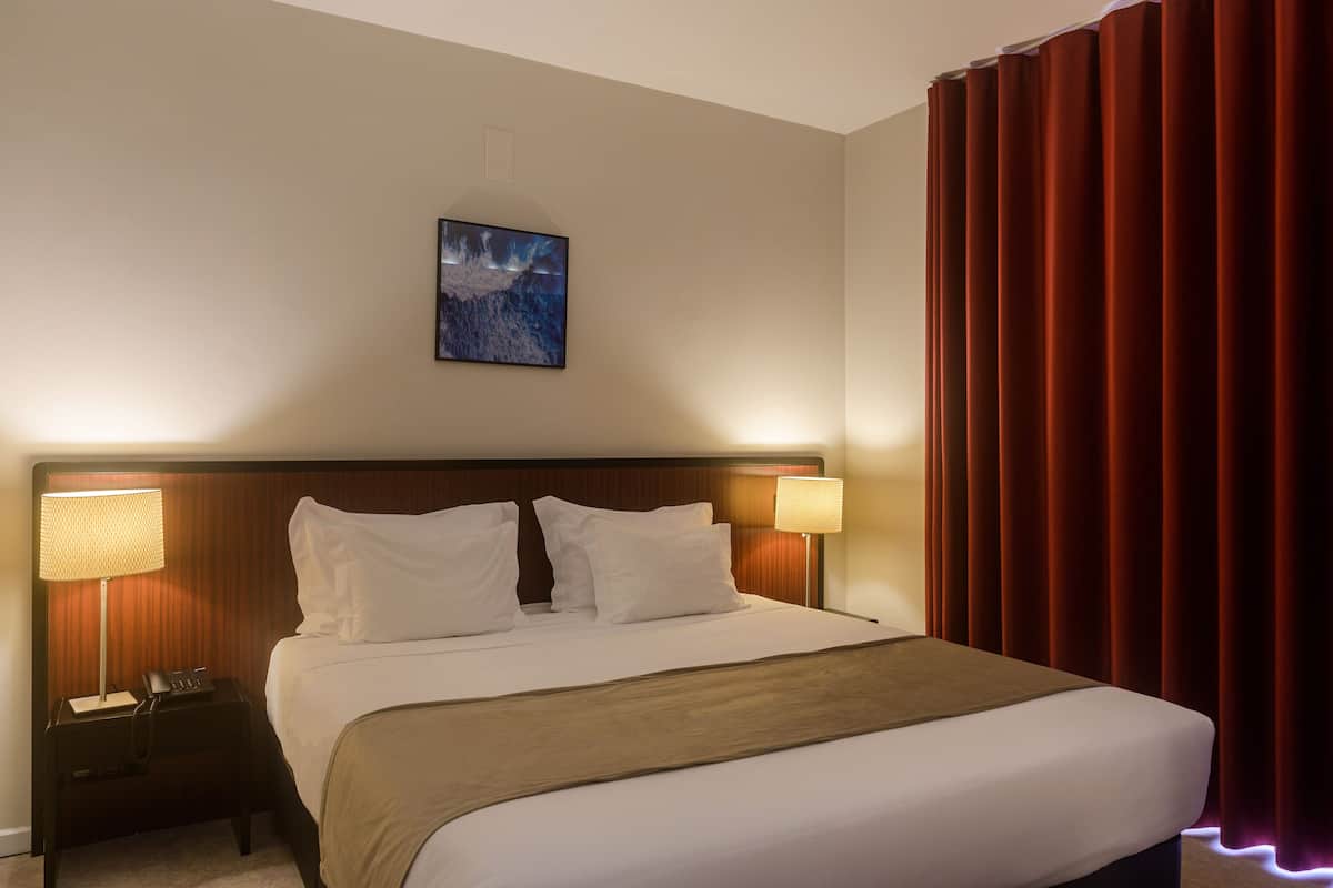 A hotel room with a large bed, two bedside lamps, a nightstand, a framed picture on the wall, and red curtains.