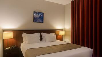 Habitación de hotel con una cama grande, dos lámparas de noche, una mesita de noche, un cuadro enmarcado en la pared y cortinas rojas.