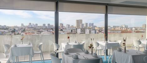 Uma área de refeições no terraço com mesas preparadas para o serviço, grandes janelas com vista para o horizonte da cidade.